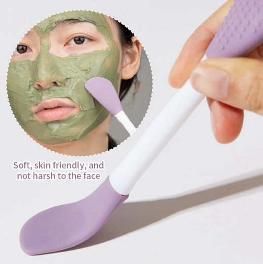 Person applying a green face mask with a double-headed silicone face brush, with an inset showing a close-up of the mask on the face.