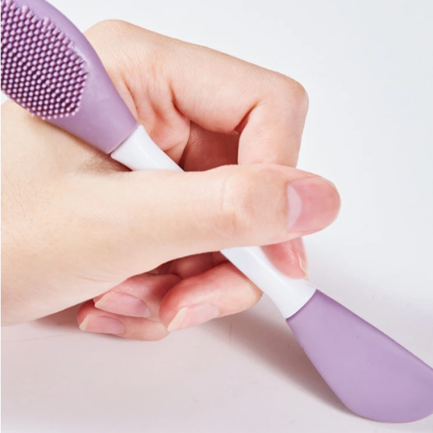 Hand holding a purple and white double-headed silicone face brush on a light background